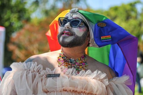 A drag queen nun with a rainbow habit, white face paint, beard, sunglasses, colorful necklace, rainbow pin that says "celebrate diversity" and a name tag that says "sister splenda derriere"