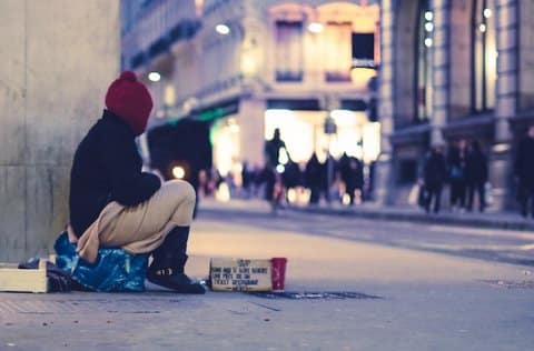 A person sitting on a city sidewalk wearing a red knitted cap and looking away from the camera