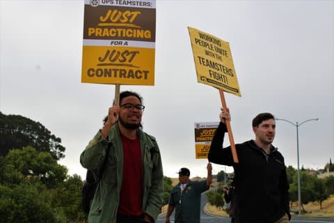 A Black man holding a picket sign that reads "UPS teamsters just practicing for a just contract" next to a white man holding a picket sign that reads "working people unite with the teamsters fight"