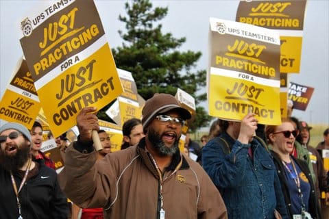 A Black man in a UPS jacket holding a sign that reads "UPS Teamsters just practicing for a just contract" at the center of a crowd of people holding the same signs