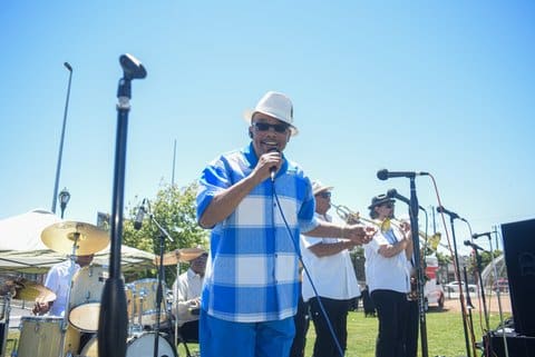 A Black man wearing a blue and white shirt, white fedora and dark sunglasses on stage on a sunny day with horn players and a drummer looks toward the camera with a microphone