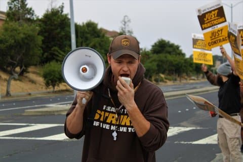A white man holding a megaphone and wearing a UPS hat, whistle and brown sweatshirt that says "ready to strike if we have to"