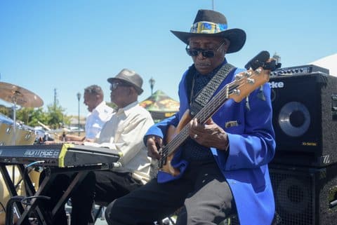 An older Black man plays the bass while seated. He is wearing a blue suit jacket and a wide-brimmed black hat with Golden State Warriors design on the band. A keyboardist and drummer, both Black men, are also visible