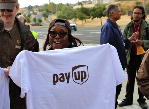 A smiling Black woman holding a white T-shirt with the message "pay up." The word "up" is inscribed in a shield design made to look like the UPS logo