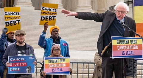Senator Bernie Sanders speaking in front of the Supreme Court building at a lectern with a sign that says "student debt cancellation is legal" along with four people with signs that say that or "cancel student debt."