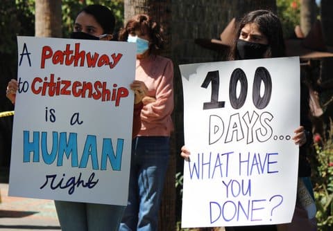 Signs that read "a pathway to citizenship is a human right" and "100 days ... what have you done?" each held by a person wearing a face mask