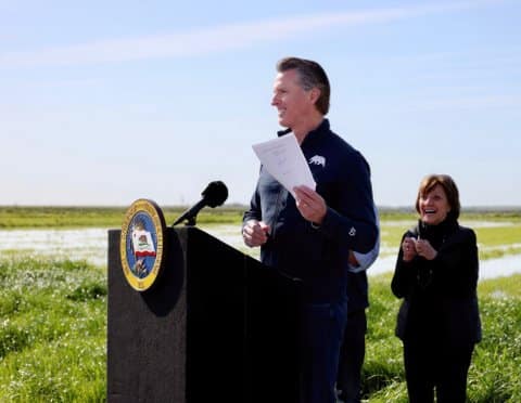 Governor Gavin Newsom holding up some paper as he stands in a field with lush grass at a lectern bearing the seal of the governor of California. A woman is smiling behind him.