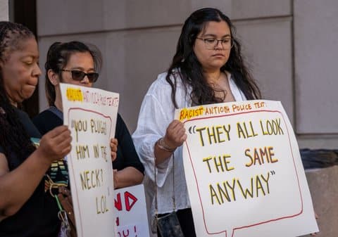 Two women of color hold up signs labeled racist Antioch police text with quotes written inside text message style bubbles One reads, "you plugged him in the neck? LOL." The other says, "they all look the same anyway." Another person is standing between them.