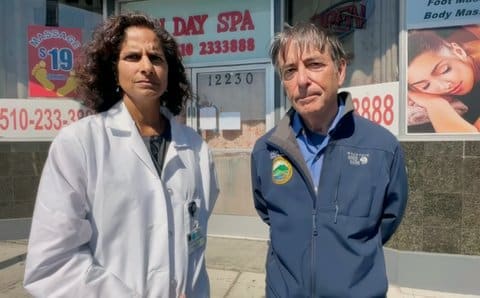 A South Asian woman in a white lab coat and a white man wearing a jacket with the Contra Costa County logo standing in front of a closed day spa