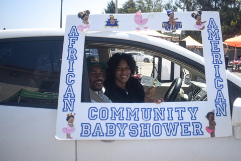 A Black man and woman in a car with a border around the front passenger window that says African American Community Baby Shower with illustrations of little Black girls in pink dresses, a pink baby carriage, a Black baby wearing a crown and sitting on a blue pillow and a Black baby wearing a hat and sitting on letter blocks