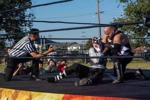 Two wrestlers compete as a referee in a white and black shirt looks on from one knee. One wrestler is wearing a white, red and black canine-style mask, silver shirt and black pants. The other is bald, unmasked, very muscular and wearing a sleeveless black shirt, black pants and tall lace-up black and gray boots
