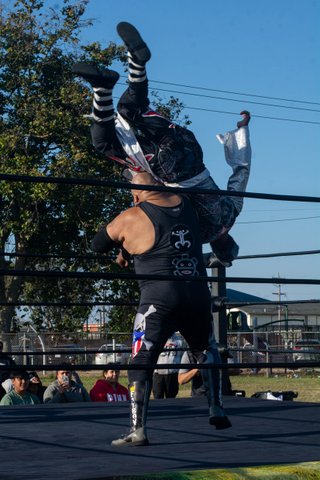 A large man in a sleeveless black top and black pants with a large skull and crossbones holds another man upside-down in the air in an outdoor wrestling ring