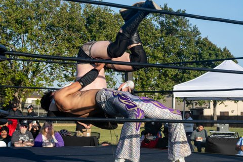 One wrestler, who is shirtless and wearing silver pants, stands bent back at the knees with his back almost parallel to the ground while another bare-chested wrestler is upside down in the air.