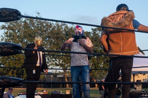 Two men in wrestling masks stand in the ring with a third man standing with his back to the camera. One of the man is dressed in a suit and tie.