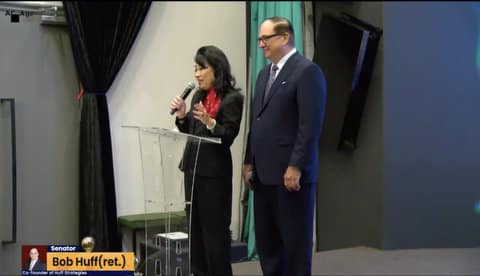 An Asian woman holding a microphone at a clear lectern with a white man standing beside her. A chyron identifies the man as retired state senator Bob Huff, co-founder of Huff Strategies.