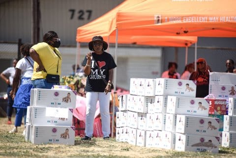 A Black woman wearing a T-shirt that says I heart my church and a Black woman in a mask standing outside among many boxes of diapers