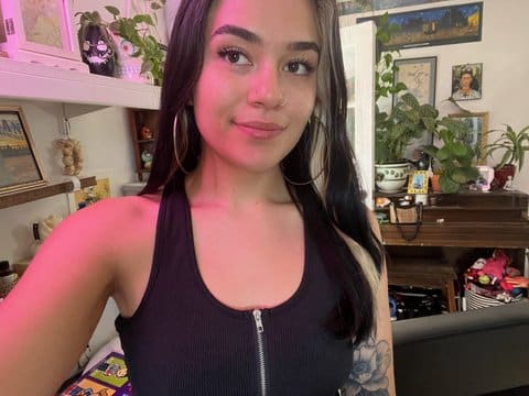 A selfie of fair-skinned young woman who is wearing a zip-front black tank top and has a rose tattoo on her left arm. She appears to be in her home, and there are plants and artwork behind her, including a painting of Frida Kahlo.