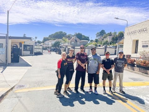 Two women and four men standing on a small road with some buildings around them. One says U.S. Post Office San Quentin California on it.