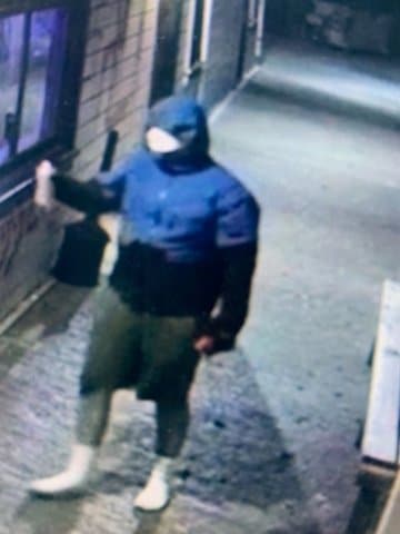 Surveillance camera footage of a person who appears to be male wearing a blue jacket with the hood up and shorts holding a can of spray paint. Their face is obscured.