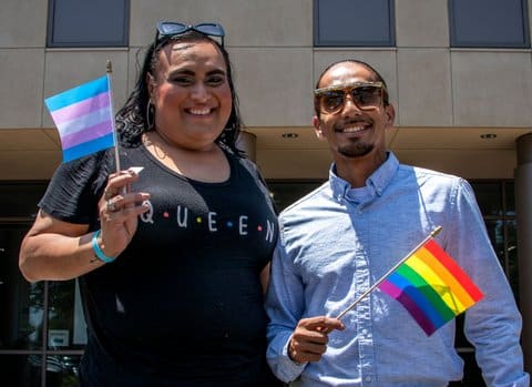 A Latina transgender woman holding a transgender pride flag and wearing a T-shirt that says queen and a Black man holding a traditional gay pride flag.