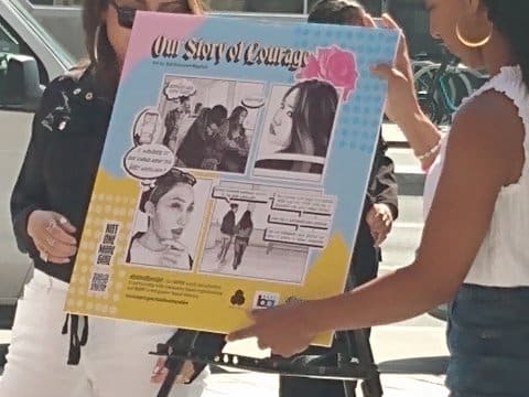 Two women holding poster titled Our Story of Courage with illustrations of women, including some on Bart train