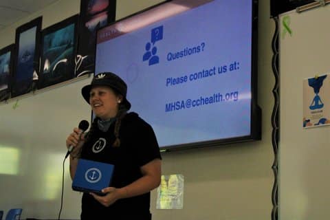 Woman wearing a T-shirt and holding a box that both have an anchor symbol in front of a screen that says questions? please contact us at MHSA at CC health dot org
