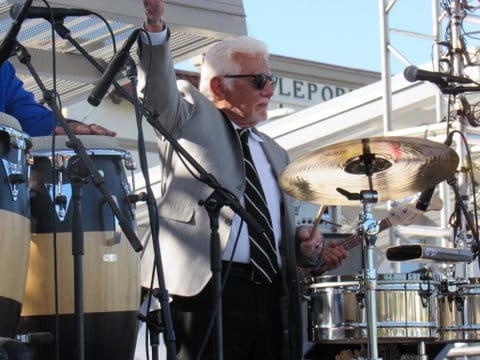 Pete Escovedo in a suit standing by his drums with one arm raised