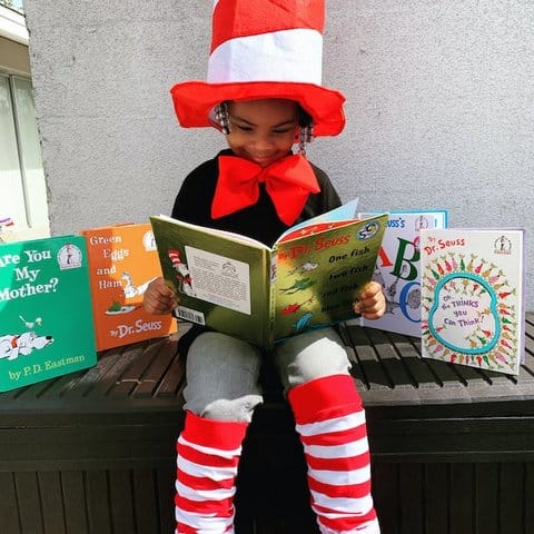 A little Black girl wearing a hat like the Cat in the Hat wears, high socks that are also striped red and white and a red bow. She is reading one Dr. Seuss book and sitting in front of others.