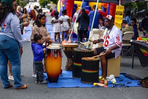 A black man wearing San Francisco 49ers cap and jersey plays African-style drums with two children playing other drums.