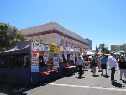 People walking outdoors past booth with signs for foods including garlic fries, funnel cake, chicken strips and wings, catfish, tilapia, fried plantains, frozen and strawberry lemonade, and corn dogs