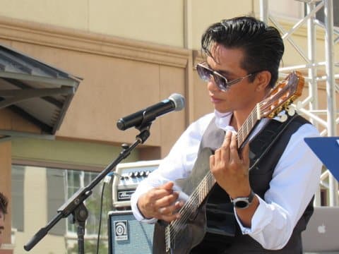 A young man in sunglasses, dress shirt and vest playing guitar