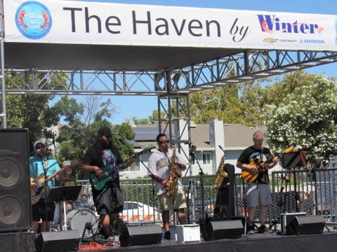 Musicians on stage playing saxophone, bass and guitar. A sign above says The Haven by Winter with logos for the Pittsburg Seafood and music festival, Honda and Chevrolet.