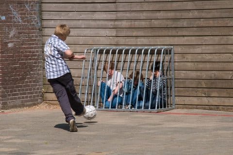 A boy kicking a ball toward a bike rack pushed against a wall with three other boys behind it like they're in a cage
