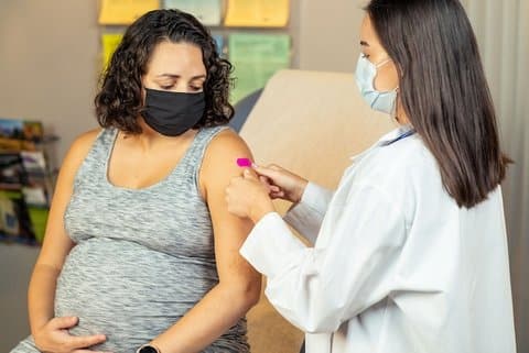 A pregnant woman with her hand on her belly looks at her arm where a woman in a white coat is applying a pink bandage. Both are wearing masks.