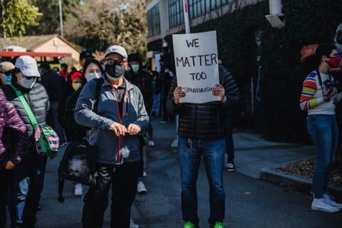 A group of Asian people in masks. One person is holding a sign in front of them that reads we matter too stand for asians.