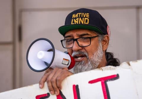 A man who is wearing a hat that says "native land" is speaking into a small bull horn