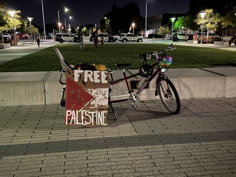A board painted with the words "free palestine" and a red triangle and white stripe similar to features of the palestinian flag. The sign is leaning against a tandem bike with a child seat in the back