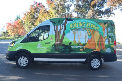 A colorful van painted with a banner that reads Contra Costa County Library Rolling Reader, along with a woodland scene with trees, grass and other greenery and a few mushrooms