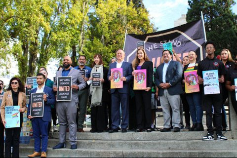 Several people of different ages, races and genders holding posters and standing on steps in front of trees. There are different posters each with a message such as "bay area stands united against hate," "protect our chosen family" (features a pride flag) and "we stand up for each other" (with a white hand and black hand reaching toward each other).