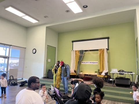 Two people speaking to an audience in front of a curtained backdrop that says rich city kick back. Five small children are standing around in the audience.
