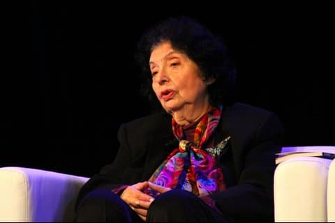 An older Jewish woman with short, dark hair wearing a colorful scarf and sitting in a white chair