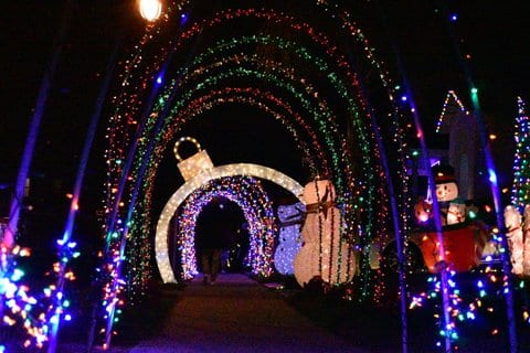 Large outdoor Christmas decoration display featuring archways covered with Christmas lights, including one shaped like a tree ornament, as well as inflatable snowmen