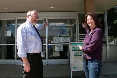 A white man and white woman standing across from each other looking at the camera both with an arm extended toward the richmond main library entrance